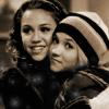 Two people in close proximity, one wearing a black leather jacket and the other in a striped beanie and dark jacket, both facing slightly right with neutral expressions.