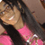 A person with long dark hair and glasses wears a pink top, smiling broadly in a close-up view.
