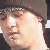 Pixelated close-up of a person's face wearing a dark cap with a light patterned band, showing partial facial features and a bright light reflection on the cheek.