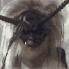 A person with long blonde hair wears a black mask and dark clothing, transitioning from masked to unmasked states