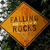 A diamond-shaped orange sign with brown text reading 'FALLING ROCKS' against a blurred background of green foliage and white light patches.