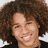 Pixelated close-up of a person with voluminous curly brown hair, light skin tone, and visible facial features including eyes, nose, mouth showing teeth in a smile.