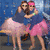 Two women in blue tops and pink skirts hug against a blue background with white speckles, transitioning from standing to crouching positions.