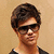 A person with short brown hair and dark sunglasses wears a black shirt with a white collar against a light brown background.
