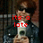 A person with brown hair and sunglasses holds a white cup, wearing a dark jacket over a light shirt against a blurred background.