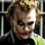 A pixelated image of a character with messy light brown hair, white face paint, dark eye makeup, and a red smile, wearing a black jacket over a green shirt against a background