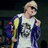 A person with blonde hair and sunglasses wears a white graphic t-shirt under an open blue jacket, holding a yellow object against a dark background.