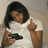 A person with dark hair wears a white shirt and holds a black handgun while pointing upward with their right hand.