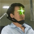 Pixelated image of a person with dark hair and beard wearing a light blue shirt, green light effect on right eye, background with white and gray tones.