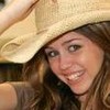 A person with long brown hair wears a light-colored wide-brimmed hat, smiling with visible teeth while holding the hat's brim with their right hand.