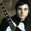 A person in a black jacket and white shirt holds a guitar with a dark fretboard and white binding against a brown textured background.