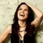 A woman with long dark hair laughs while raising her right hand to her head, set against a light textured background.
