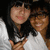 Two people in white shirts pose closely together, one with dark hair and bangs making a hand gesture near the chest.