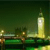 Pixelated nighttime cityscape with a tall illuminated tower, bridge, and water reflections under dark green sky.
