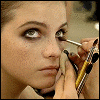 A close-up of a person applying makeup with a brush near their eye, holding a hand to their face, and holding multiple makeup tools.