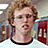 Pixelated image of a person with curly light brown hair wearing a white shirt with black collar, set against blurred background with bookshelves and red objects.