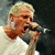 A person with short blonde hair sings into a microphone, wearing a white shirt against a dark background.