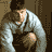 Pixelated static image of a person in light-colored clothing leaning forward with hands near the face, set against a dark brown background.