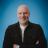 Pixelated static image of a bald man in a black jacket over a white shirt against a blue background.