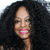 Pixelated close-up of a person with voluminous dark curly hair, light skin tone, and pink lips smiling against a dark background.