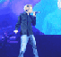 A person in a black jacket and light blue jeans sings into a microphone on stage with blue and purple lighting.