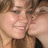 Two people in close proximity with one person kissing the other's cheek, both wearing dark clothing.