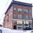 Pixelated static image of a building facade with multiple windows and a dark entrance, rendered in muted brown, blue, and gray tones.