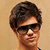 Pixelated static image of a person with short dark hair, wearing black sunglasses and a light-colored shirt against a beige background.