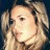 Pixelated close-up of a person with light brown hair styled in two braids, wearing dark clothing against a dark background.