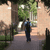 Pixelated static image of a person walking on a path between trees with green foliage and light-colored buildings in the background.