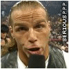 A person with short light brown hair wearing a dark vest over a white shirt speaks into a black microphone against a blurred crowd background.