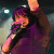 A pixelated figure with purple hair holds a microphone, wearing dark clothing against a blurred background with light effects.