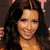 A woman with long dark wavy hair and light skin smiles at the camera, wearing a strapless top against a blurred background.