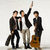 Three pixelated figures in dark suits stand against a light background; one raises an arm, another holds a guitar-like object.