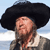 A pixelated figure with a large black hat, long brown hair, and a light-colored shirt against a blue sky background.