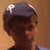 Pixelated close-up of a person's head and neck wearing a dark cap with light reflections, set against a warm brown background.