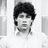 A grayscale pixelated portrait of a person with dark curly hair, wearing a white shirt against a light background.