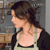 Pixelated image of a person with dark hair wearing a light-colored tank top and dark cardigan, facing left against a blurred background.