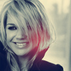 A static portrait of a person with short light-colored hair, smiling, wearing a dark top against a blurred background with red and blue color grading.