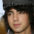 Pixelated close-up of a person with dark hair and light skin, wearing a dark top against a blurred background with visible text.