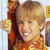 A young person with light brown hair and bangs smiles broadly, wearing a colorful patterned shirt with orange and yellow tones.