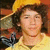 A person with curly brown hair wears a yellow cap and shirt against an orange background, with text appearing in the lower right.