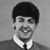 Black-and-white pixelated image of a person with short dark hair, wearing a white collared shirt and dark jacket, smiling with visible teeth.