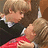 Two figures in close embrace, one with light brown hair wearing dark clothing and the other with blonde hair in a red garment.
