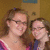 Two women in a school hallway pose together, one wearing glasses and pink-and-purple clothing with a necklace, the other in dark blue.