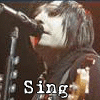 A person with shoulder-length dark hair sings into a microphone, wearing a dark jacket over a light shirt. Text and vertical bars change across frames.