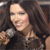 A pixelated close-up of a person with long dark hair holding a silver microphone, wearing a dark top with light accents.