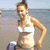 A woman in a white bikini top with green accents stands on a beach, her right hand on her hip and left arm bent at the elbow.