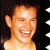 Pixelated close-up of a smiling person with short light brown hair, visible ear, and dark background with white star-like shapes.