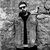Black-and-white pixelated image of a tall, thin figure with a rounded head and dark vertical body against a textured background.